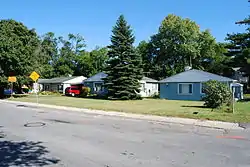 Three small one-story houses with pointed roofs on a street with tall trees behind them