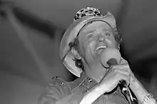 A black and white photograph of a smiling man wearing a cowboy hat and holding a microphone