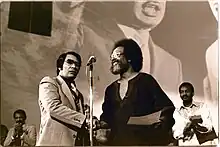 Jim Jones shakes hands with Cecil Williams with a large picture of Martin Luther King Jr. in the background.