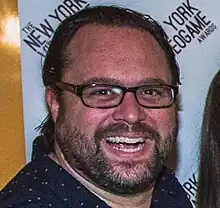 A 42-year-old man with black hair, a black beard, and glasses smiling with an open mouth at the camera.