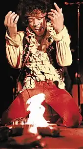 A color photograph of a man kneeling over a guitar that is on fire