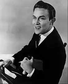 A dark-haired young man wearing a dark jacket and a necktie, smiling broadly while sitting in a chair and turning to look behind him