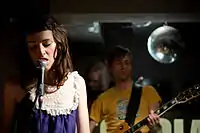 A brunette woman singing into a microphone. In the background there is a male guitarist wearing a yellow t-shirt, and a glitter ball