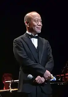 Joe Hisaishi standing on stage in Paris in 2011.  He is standing with his wrists crossed with a microphone in hand and a set of index cards in the other.  He is smiling.