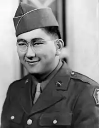 Head and shoulders of a smiling young man with dimples and round wire-framed glasses wearing a garrison cap and a military jacket over a shirt and tie.