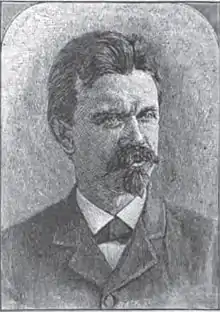 A man with thick, dark hair, a mustache and a goatee, wearing a white shirt and dark jacket