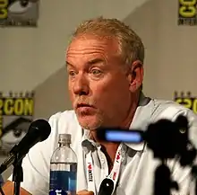 John Debney speaking at ComicCon 2013.  He is sitting at a table, with a microphone in front of him and a white backsplash with logos behind him. He is speaking, and in the middle of a hand gesture.