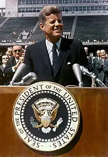 Kennedy, in a blue suit and tie, speaks at a wooden podium bearing the seal of the President of the United States. Vice President Lyndon Johnson and other dignitaries stand behind him.