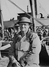 A middle-aged man posing for a photo wearing a World War II-era military uniform. The superstructure of a ship and the bow of a boat are visible behind him