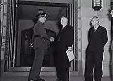 Black and white photograph of a man wearing military uniform shaking the hand of a man in a formal suit. The two men are standing in front of a stone gateway, and another man wearing a formal suit is looking on from the right of the image.