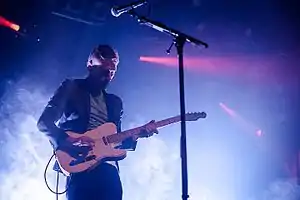 Johnny Blake plays his guitar while standing in front of a microphone. A bright blue light shines on Johnny, and other spotlights in the background aim elsewhere