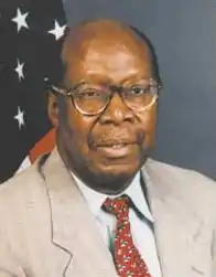 A portrait photo of a balding black man in front of a US flag; he is wearing spectacles, a tan suit, red tie, and is looking to the camera's right.