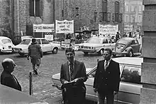 In the foreground Van Agt enters a building on the Binnenhof in The Hague. In the background there are about ten demonstrators behind crowd barriers. Banners read: "Stop this child murder", "Both left and right enforce current law!!" and “If you want peace, defend life.”