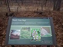 Large brown sign with pictures of blue butterflies next to a trail through oak savanna