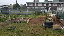 Depicts garden beds in the foreground and some seating, with some houses and a small apartment building across the street. Sunny day with blue sky.