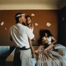 In a bedroom, Kendrick Lamar wearing woven crown of thorns is seen holding an infant child with a woman seen sitting on a bed holding another baby. A gun can be seen at the waistband at his back. The bedroom has brown paint with white spots and a radiator.