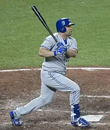 A man, wearing a blue batting helmet and grey baseball uniform, swings his bat