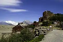 alt=Photograph of the red-painted Kennecott Mines facilities rising up a hillside with grand mountain scenery in the far background and a field of bare glacial till in the middle background.