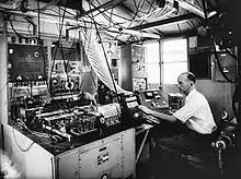 Monochrome photograph of man in room with electronic equipment and many cables straggling from the ceiling and elsewhere, with his hand on a black machine.