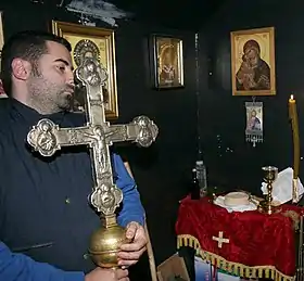 Photograph of a man in his thirties holding a large silvery cross in a room on whose walls religious paintings are hung.