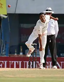 A man in Test cricket whites, on his follow through after releasing the ball while bowling; three stumps and a man in white pants, white shirt and hat are in the background.