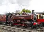 A black pannier tank locomotive is travelling from right to left, bunker first, along a single track on the side of a wooded hill. The locomotive is pulling five passenger carriages painted cream and brown, and a plume of smoke is passing over the first two carriages. The image of the train is reflected the river in front of the train.