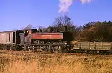 A pannier tank locomotive is pushing a shunter's wagon and pulling a guard's van. The locomotive is painted in a faded maroon with black and yellow piping, but much of the paint has peeled, revealing the black paint underneath. The lettering "LONDON TRANSPORT" is shown in yellow on the side of the pannier tank.