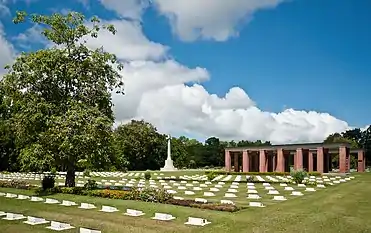 Colour photo of a cemetery comprising rows of small stone grave markers. A cross-shaped monument and a red brick structure are visible in the background