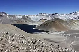 A dark lake in a fairly deep crater; the terrain is in part covered by snow, in part rocky, with no vegetation