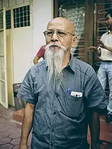 Mong Hay standing in a street. He is bald with a long white bear and wearing a collared, short sleeved shirt with a pen and smart phone in the pocket.