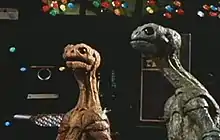 Two reptile-like alien creatures, one blue and one orange, look in an upward direction, with machinery and computer screens in the background.