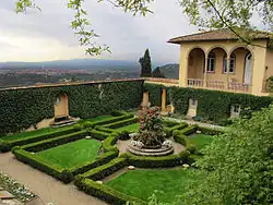 A yellow stucco building with a red clay roof and several arched doors and windows, surrounded by green shrubs.