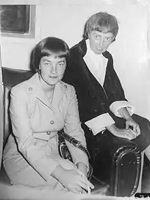 Photograph of two short-haired young women wearing suits and sitting in an arm chair. The woman in the chair has a cigarette in her hand and the woman sitting on the arm of the has her hands clasped in her lap.