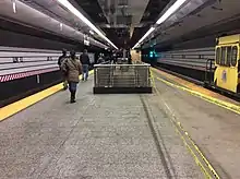 The Lexington Avenue–63rd Street station undergoing renovation, with new tiling and platform treads on the platform, and new walls next to the subway tracks on either side. A work train is visible on the right-hand track, which is cordoned off with yellow caution tape. In the background, passengers can be seen heading toward the escalators to exit the station.