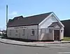 Three-quarter view of a long, low, cream-coloured painted hut with white cladding below the tiled roof. An entrance porch with a brown door extends from the shorter side; the longer side, alongside a street corner, has four rectangular windows. "The Lighthouse Community Church" is written in red letters above these.