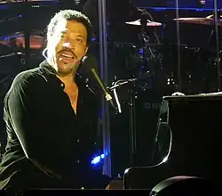 A dark-skinned man with dark hair and a thin mustache playing a piano and singing into a microphone