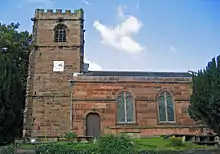 A sandstone church seen from the south; on the left is a crenellated tower with a square clock face; to the right is the body of the church with a doorway and two windows, all round-headed