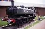 A green pannier tank locomotive stands on a short length of track on a gravel bed surrounded by grass. In the background is a brick building. The letters G W R are shown on the side of the pannier tank. A small box stands near the cab and is pointing to the wheels.