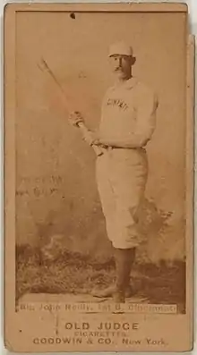 A man wearing an old-style white baseball uniform and pillbox hat