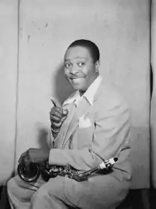 A dark-skinned man wearing a light-coloured suit, smiling broadly while holding a saxophone with one hand and pointing upward with the other