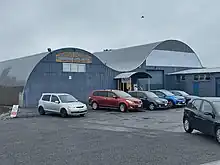 A curved building with two swoops like the letter M - single level corrigated iron building with a shed attached. Parked cars in the front, and people under the entrance roof..