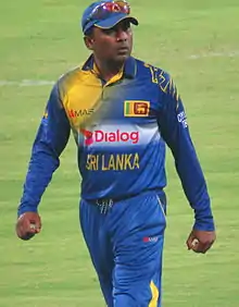 Portrait of dark skinned man, wearing blue and yellow Sri Lanka cricket team kit with cap. Cricket field in the background.