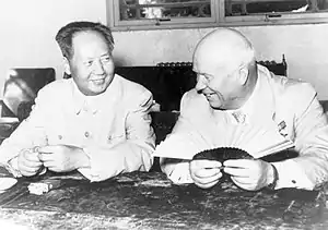 A balding man and a younger Chinese man sit and smile, the balding man holding a fan