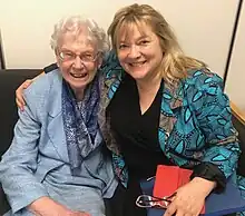 Margaret-Ann Armour (wearing glasses) is dressed in a gray suit with a scarf around her neck. On the right, Imogen Coe is wearing a green blazer and black shirt, and is holding a red clutch.