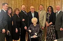 Martha Schwebach sits in the middle of her family at the 2023 awards ceremony for the New Mexico Center for Nursing Excellence. Family members, middle-aged, are dressed formally before a background of a gold sequined curtain. Martha, elderly, with white curly hair, is seated in a wheelchair, with her husband Don behind.