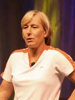 A blond-haired women with a white shirt, black shorts, and white tennis shoes on about ready to serve the tennis ball in hand