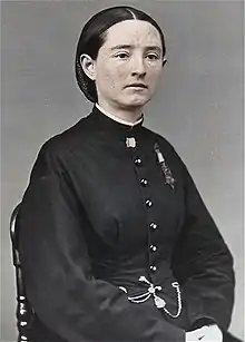 Head and shoulders of a white woman with straight hair pulled back into netting, wearing a tunic with a star-shaped medal hanging from a ribbon pinned to the left breast.