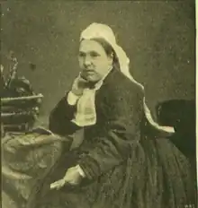 B&W portrait photo of a seated middle-aged woman in a dark dress, wearing a white head scarf and a white lace kerchief.