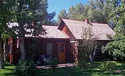 An unpainted dark brown wooden house amid tall trees in two sections. The taller one, on the right, has a pointed roof with a small brick chimney rising from the middle.