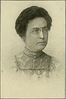 A middle-aged white woman with dark hair in an updo, wearing pince-nez glasses and a high-collared lacy dress